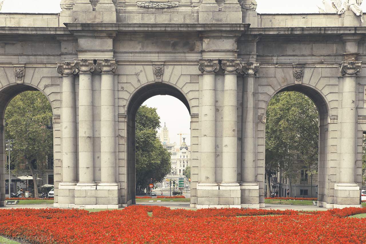 Foto von Stone archways frame city life amidst vibrant red flowers, a serene snapshot of Madrid's grandeur.