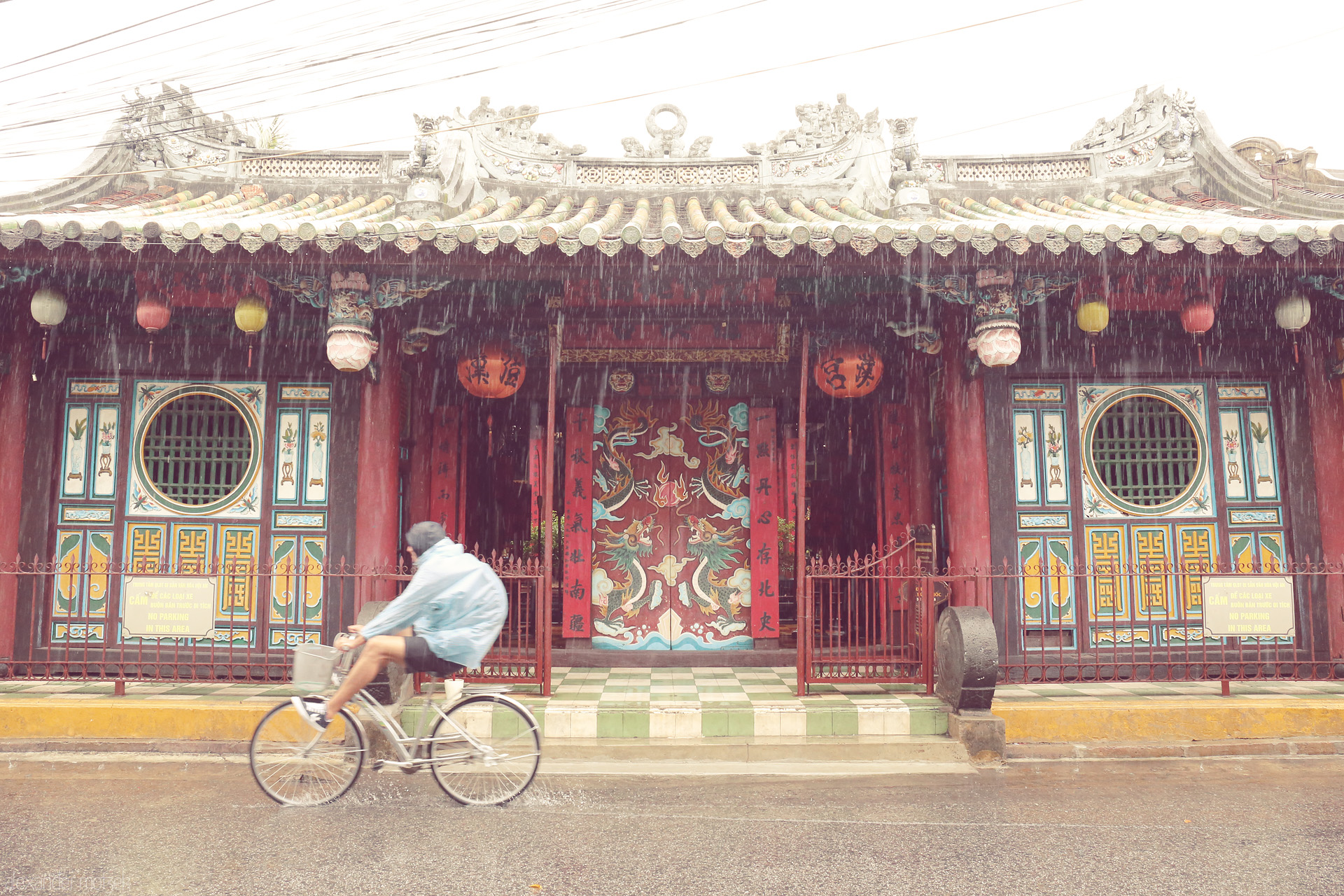 Foto von A cyclist glides past the dragon doors of a Hoi An temple, rain dancing off ancient lanterns in Vietnam’s poetic embrace.