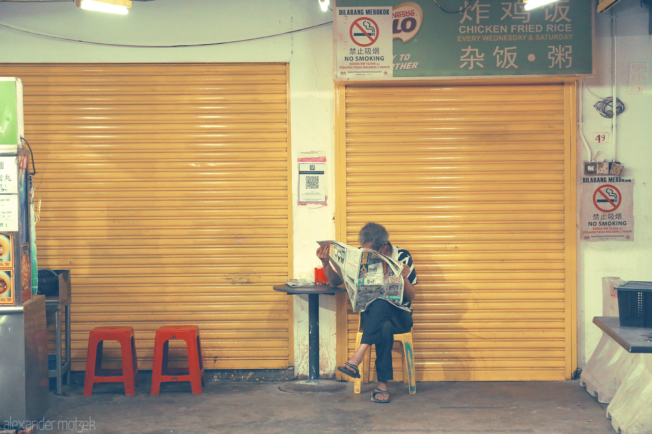Sips and Stories Foto von A local enjoys a quiet moment with a newspaper at a closed stall in Kuala Lumpur's bustling market scene.