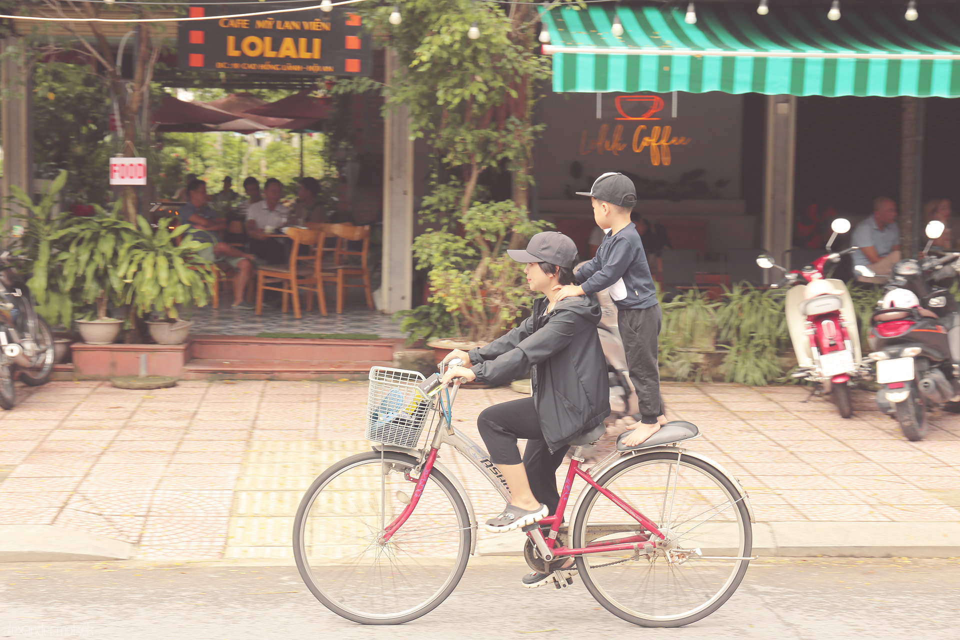 Foto von A mother and child glide by a cozy Hoi An café, balancing daily life and joy on a bicycle in Vietnam’s lantern-lit town.