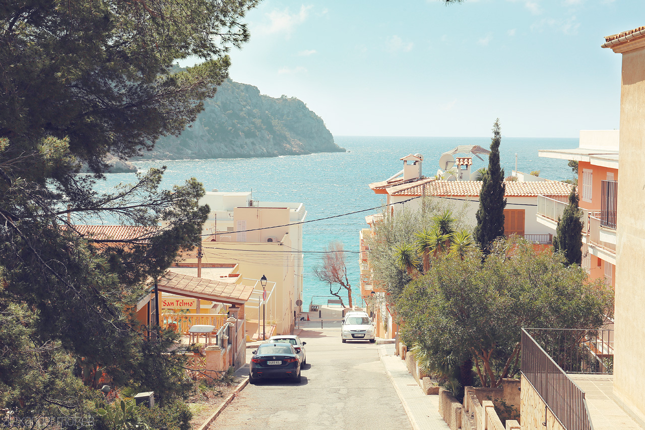 Foto von A serene glimpse down a tree-lined street in Sant Elm, Mallorca, leading to the azure Mediterranean Sea.