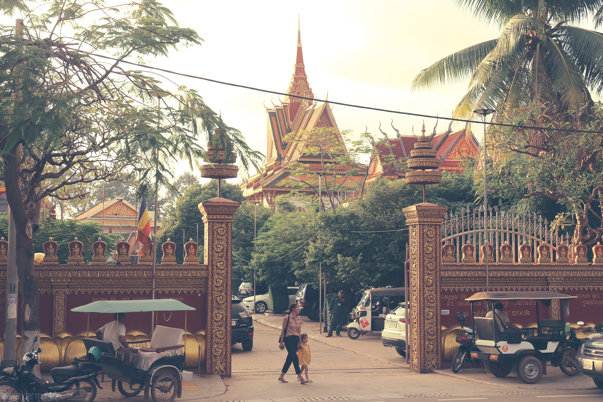 Foto von A woman and child walk past golden gates and tuk-tuks, with a Khmer temple shimmering under Siem Reap’s lush palms and late sunlight.