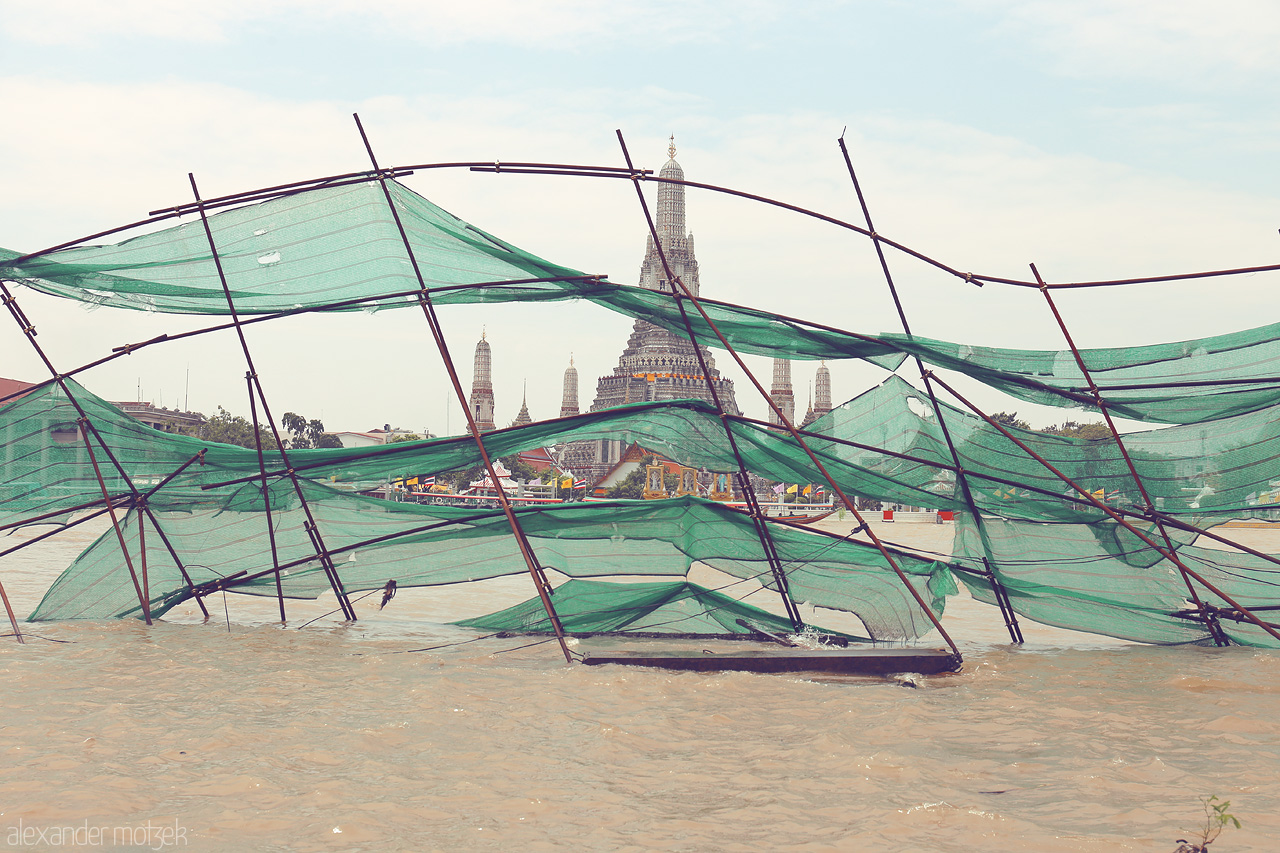 Foto von A serene view of Phra Borom Maha Ratchawang across the river, framed by delicate green nets, capturing the essence of Bangkok's timeless beauty.