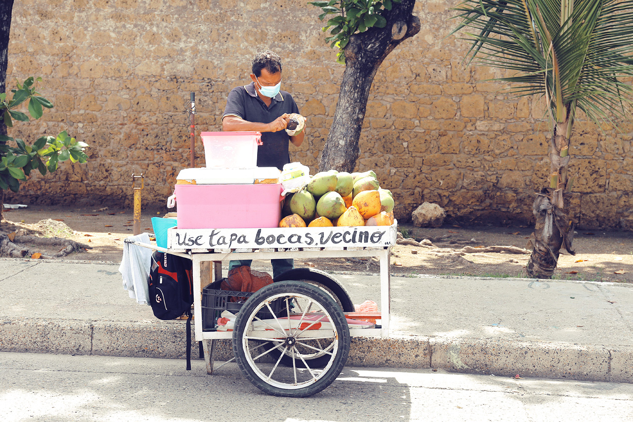 Foto von A street vendor in Cartagena, Colombia serves fresh coconuts and local treats along a sunlit, cobblestone pathway.