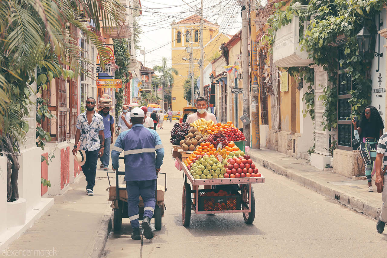 Foto von A vibrant street scene in Cartagena, Colombia featuring a fruit vendor surrounded by colorful architecture and lively people.