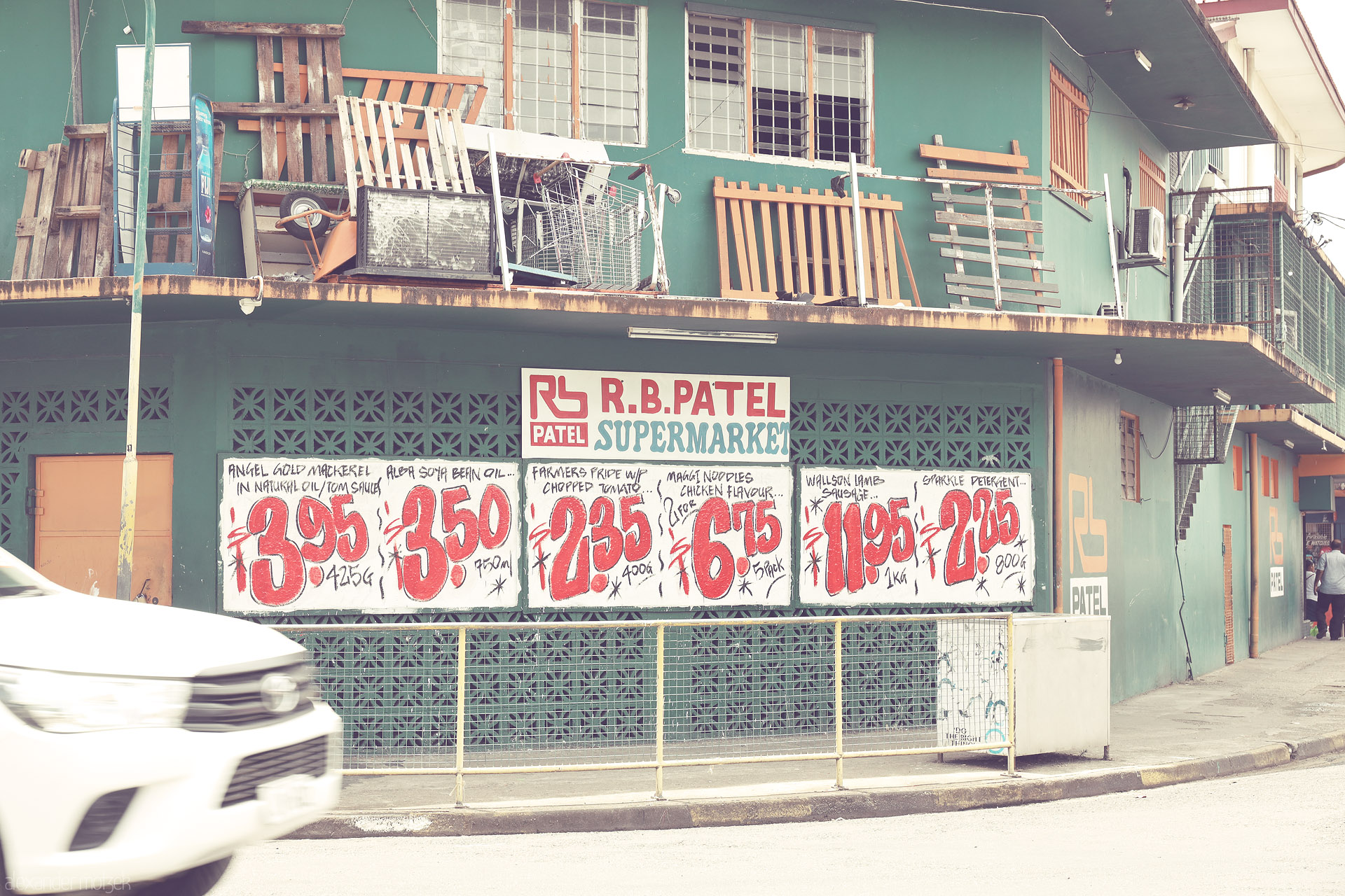 Foto von R.B. Patel Supermarket facade with hand-painted price boards and a passing car, on a street in Sigatoka, Fiji.