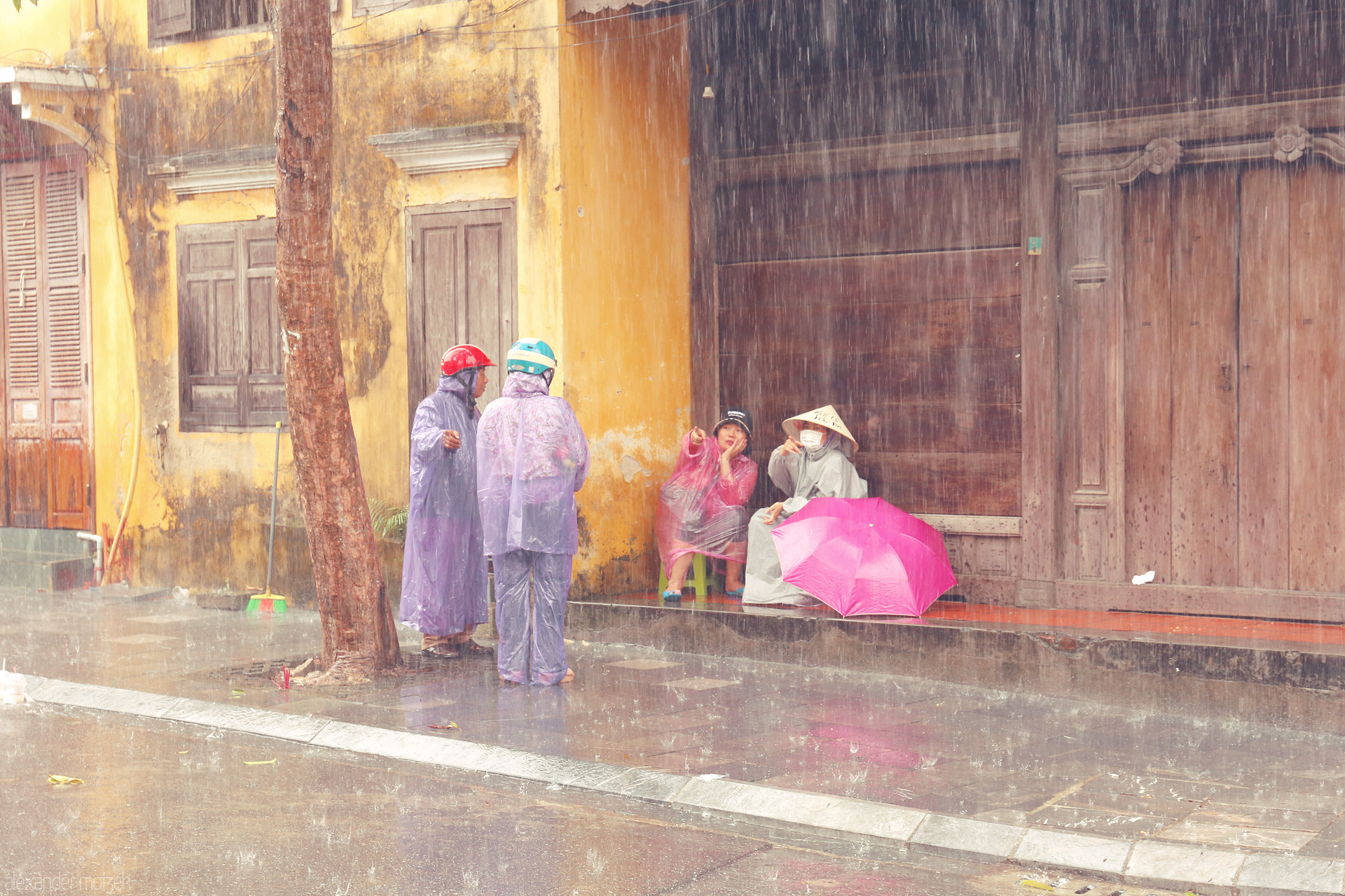 Foto von Locals pause under the rain in Hoi An, Vietnam, bright raincoats and a pink umbrella glowing against ancient yellow walls.