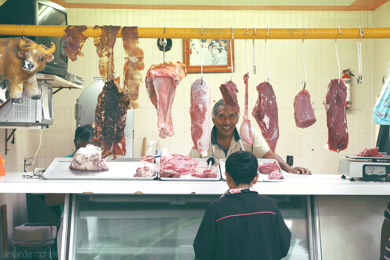 Foto von Joyful butcher in Oaxaca's market, with hanging meats and an engaged customer.