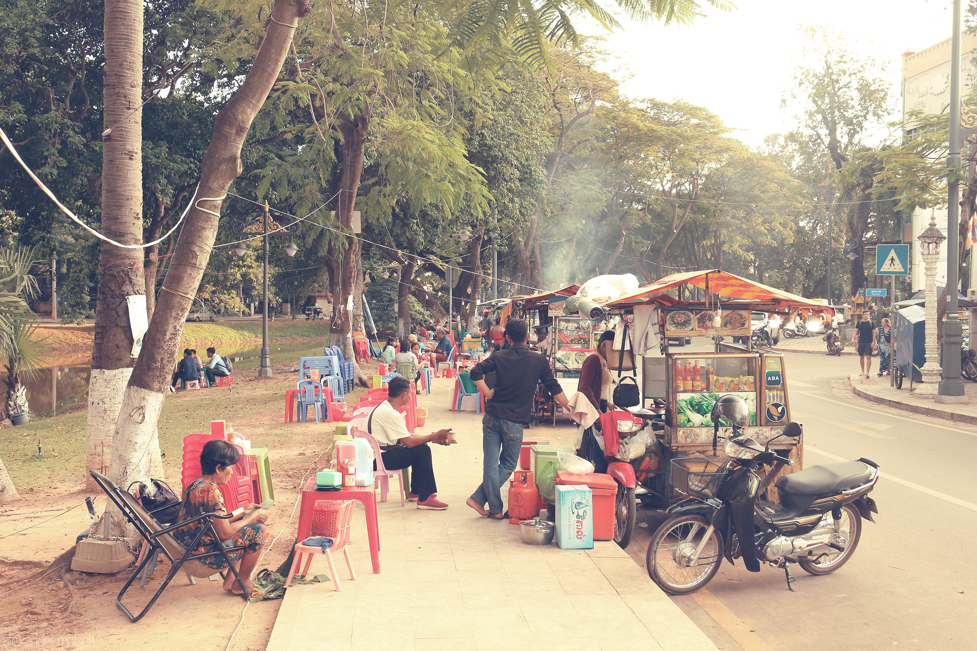 Foto von Morning bustle in Siem Reap: street food carts, plastic chairs, and locals savor moments by the riverside under lush tropical trees.