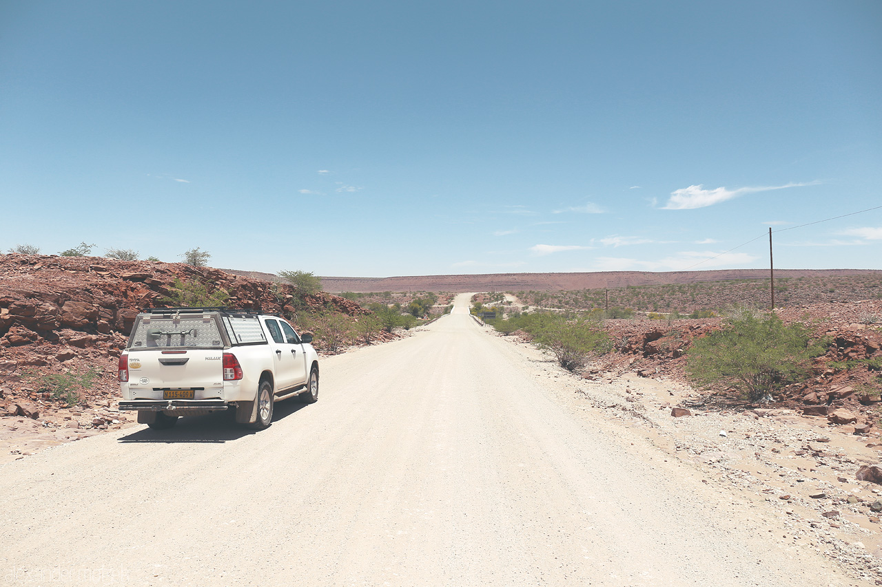 Foto von A lone vehicle traverses the gravel roads amidst Hardap's rugged terrain under the vast Namibian sky.