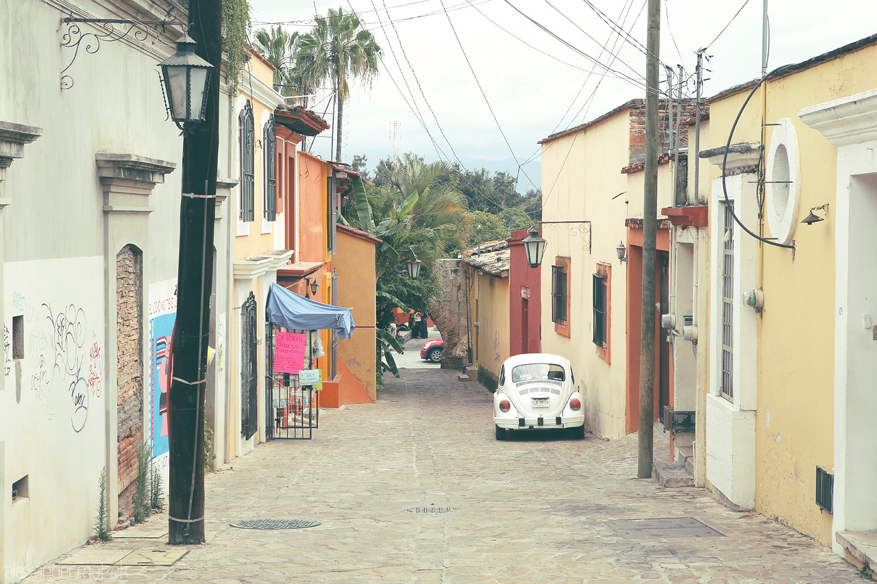 Oaxaca Callejones Foto von Narrow cobblestone street, an old off-white volkswagen beetle, and colorful facades capture the essence of Oaxaca de Juárez, Mexico.
