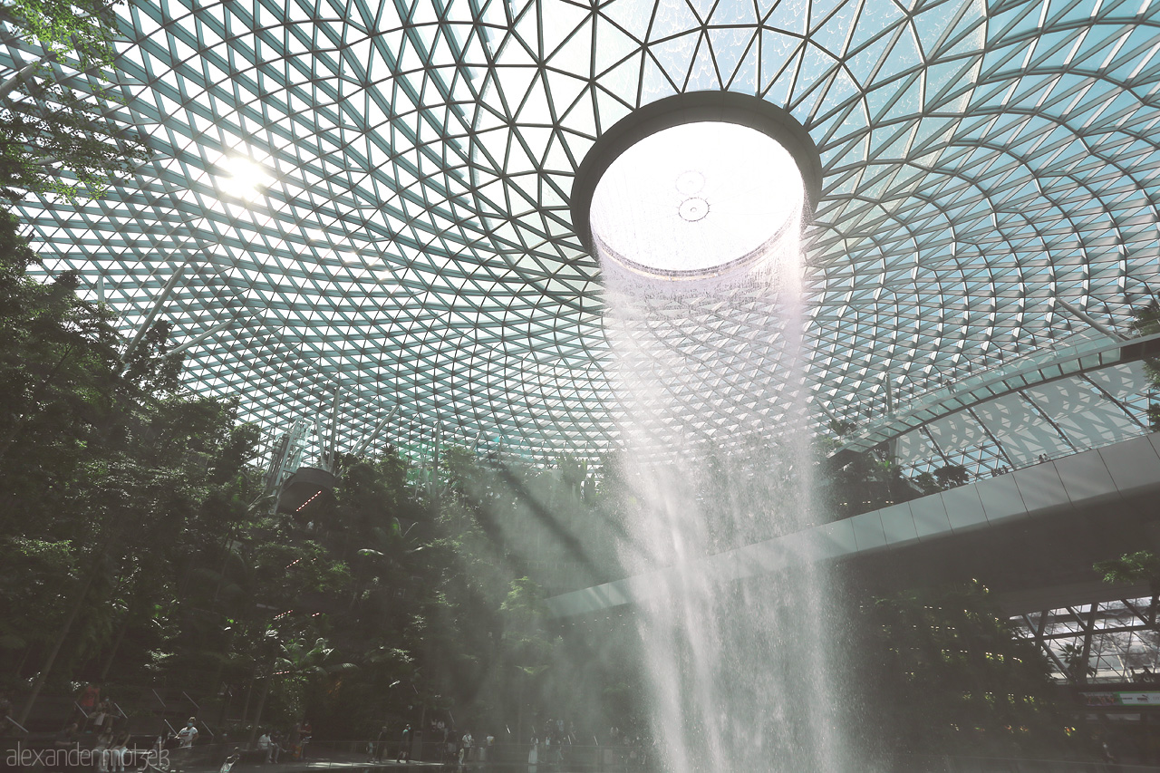 Foto von A mesmerizing waterfall beneath a vast glass dome at Jewel Changi, Singapore, surrounded by lush greenery and sunlight streaming through.