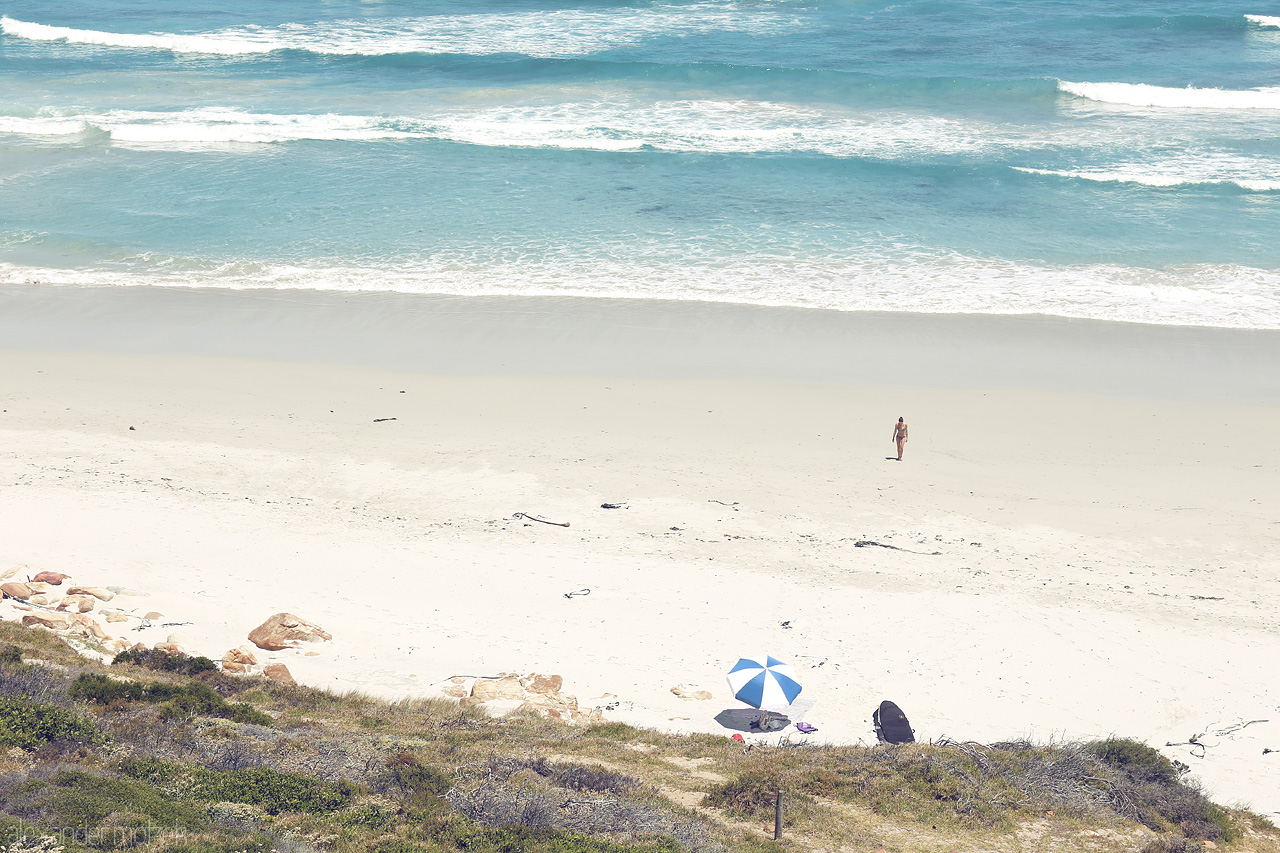 Foto von A lone figure stands on a serene Cape Town beach, waves gently lapping at the shore, with a solitary umbrella providing shade.