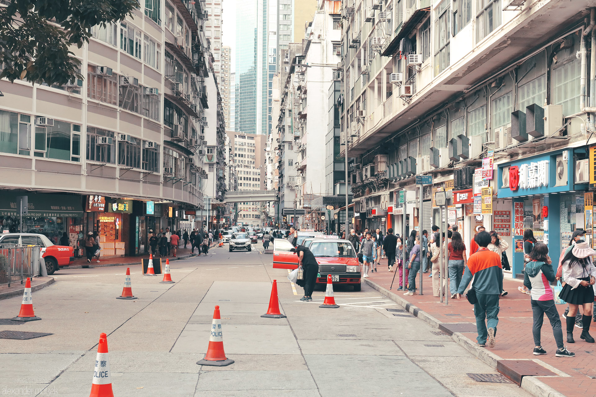 Foto von Street life in Wan Chai, Hong Kong—red taxi by orange police cones, shop signs and crowds beneath dense apartment blocks.