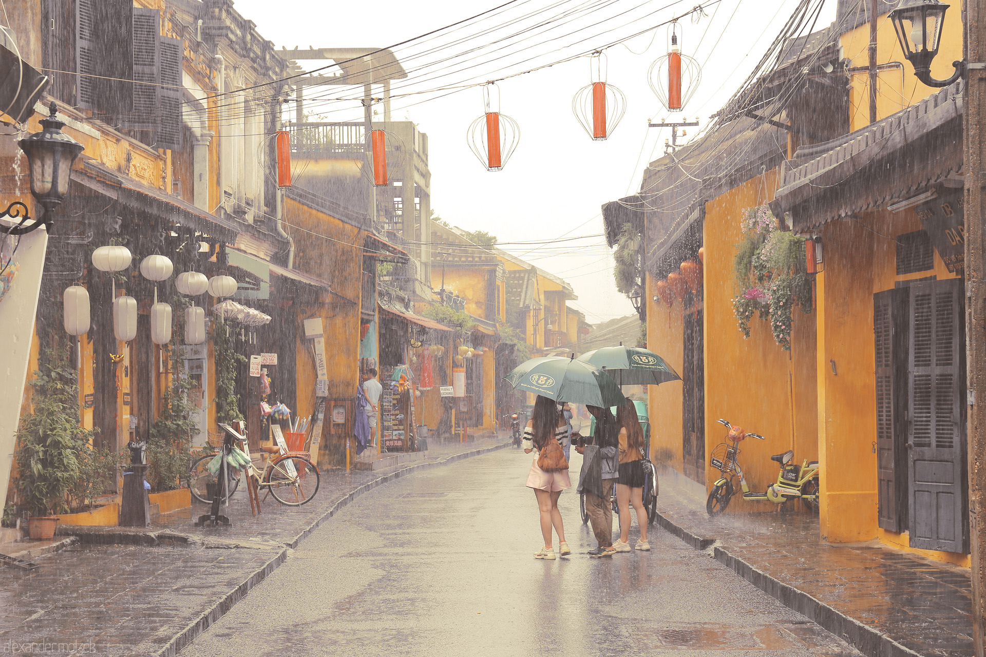 Foto von Three friends under umbrellas stroll Hoi An’s golden old town, lanterns aglow amid a gentle summer rain in Vietnam.