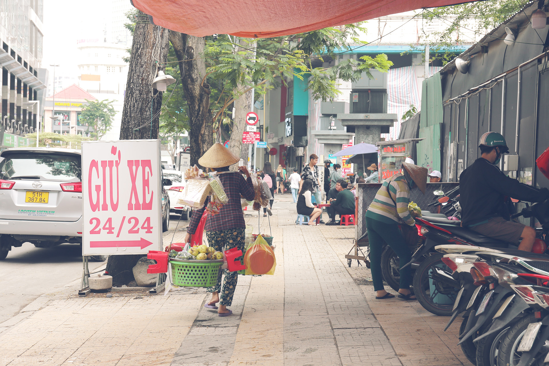 Foto von A street vendor in a nón lá weaves through Saigon’s bustling sidewalks, framed by local life, scooters, and a bold 'Giu Xe' sign.